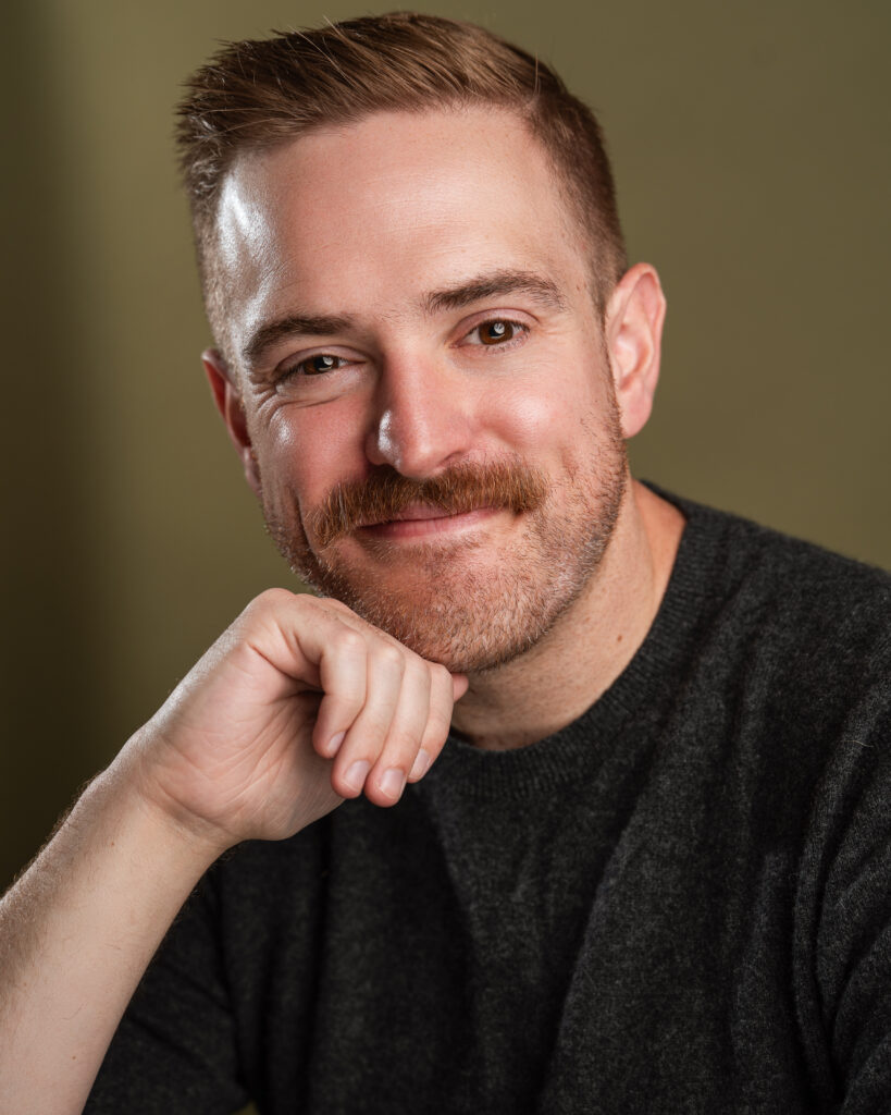 Headshot of Jake leaning against his hand. He has a soft smile.